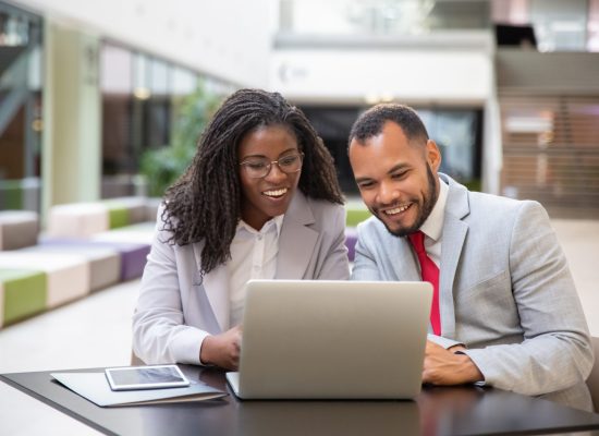 Happy excited colleagues using laptop for video call. Business man and woman sitting in office lobby, staring at computer monitor, smiling and speaking. Video chat concept
